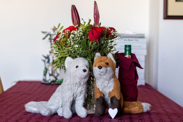 Two fox plushies sitting on a table in front of a vase with red roses.