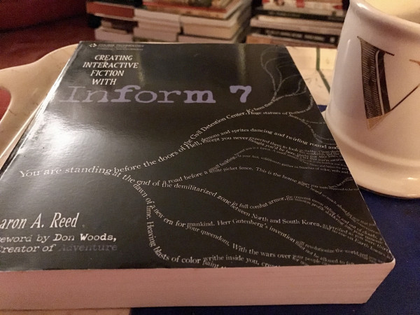 A hefty paperback copy of Aaron A Reed’s book “Creating interactive fiction with Inform 7”. The book is resting on a tray beside a big cream mug with a gold monogrammed V.