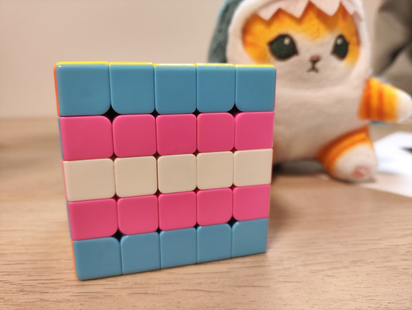 A 5x5 stickerless Rubik's cube arranged with the trans flag colors, with a cathaj plushie behind