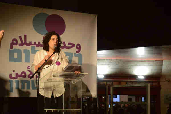 Rula Daood speaking into a microphone, standing in front of a large sign that reads “Only Peace Will Bring Security” in Arabic and Hebrew.