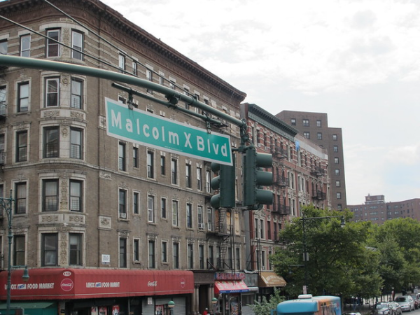 Picture of a block in NYC featuring a streetsign in the foreground reading "Malcolm X Blvd" and some apartment buildings in the background