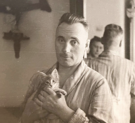 Black and white photo of a handsome white man wearing pajamas and smiling a little as he holds up a tabby kitten. Both man and kitten are looking at the camera. The photographer can be seen in the mirror behind the man.