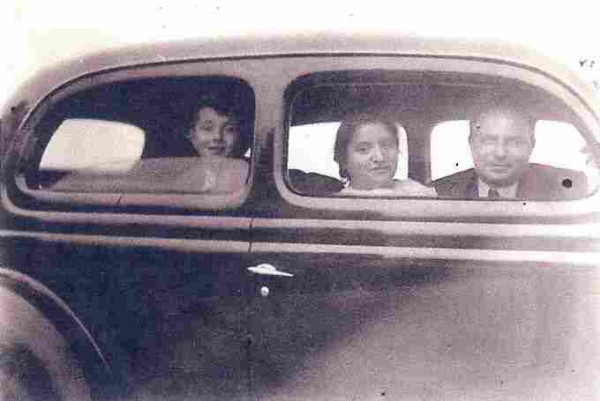 Photo of the side of an old car. The front open window shows the face of a man and a woman. In the rear window, a young boy's head can be seen from behind the open window.