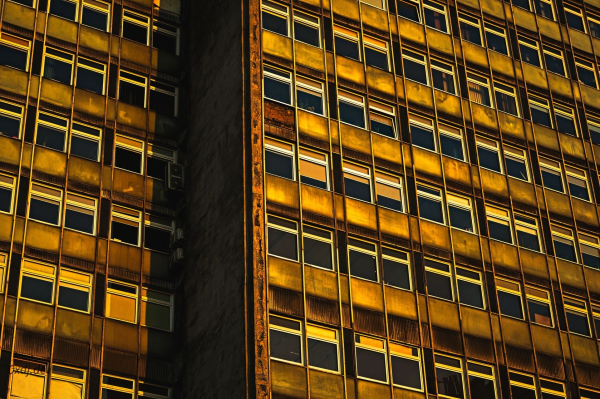 A facade of an old office building, with many identical windows, some of them partially covered by window blinds. The building has two parts, one part is set slightly back from the other, more visible in the photo. The concrete parts of the facade are visibly old and have some defects. The colour palette of the photo is warm, orange-yellow-ish, reflecting the golden hour during which it was shot.