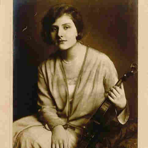 A woman photographed in a studio. She is sitting. He has long hair, reaching her neck. In her left hand, she is holding a violin. 