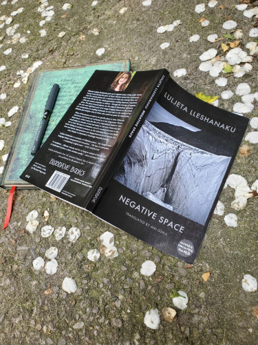 Poetry collection Negative Space by Luljeta Lleshanaku, translated by Ani Gjika (Bloodaxe Books) sits on a cement walkway with a notebook with an ornate green cover and a black pen. The walkway is covered in mock orange blossoms.