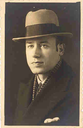 Portrait photograph of a man wearing a coat. He is elegantly dressed - a handkerchief around his neck and a handkerchief in his coat pocket. On his head is a hat with a large brim.