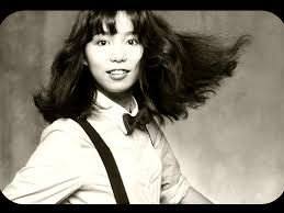 Black and white photograph of Japanese pop singer  Mariya Takeuchi. She is wearing a dress shirt style  blouse, a bow tie, and dark suspenders. Her shoulder length hair is blowing in a breeze and she looks into  the camera with a smile .