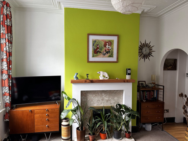 A picture of a living room, showing the chimney breast. To the left is a sideboard with a tv on top. To the right a chest of drawers with a sunburst clock above. A picture hangs on the chimney breast, it is a schools illustrated poster of Henny Penney. There is a wooden mantelpiece with a range of ornaments. The chimney breast is painted in Dulux Luscious Lime, a fresh mid to light green with plenty of warmth to it. There are houseplants on the hearth.