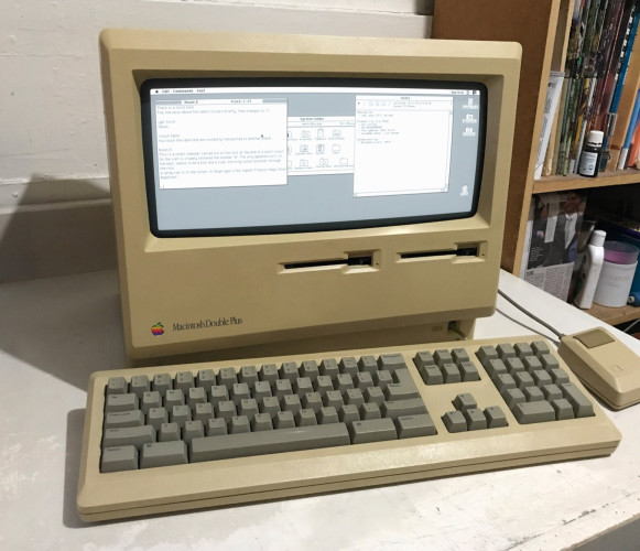 A picture of an old and yellowed Macintosh Double Plus on a desk. It's much like a regular Macintosh Plus, but has two floppy drives, and a double-width greyscale CRT. The keyboard features a nav cluster and numpad. The mouse has one button.