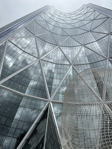 A colour photograph, April 12, 2024. Looking upward at a tall curving office building. The windows appeared to be mirrored. There are large diagonal crossing elements on the exterior of the building.