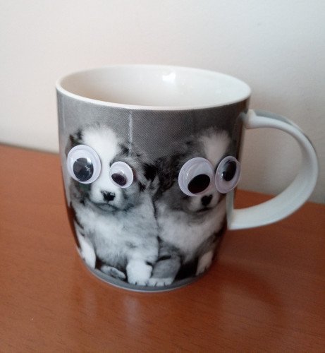Photo of a mug with two dogs on it. There are big googly eyes stuck over the normal eyes giving a comical effect 