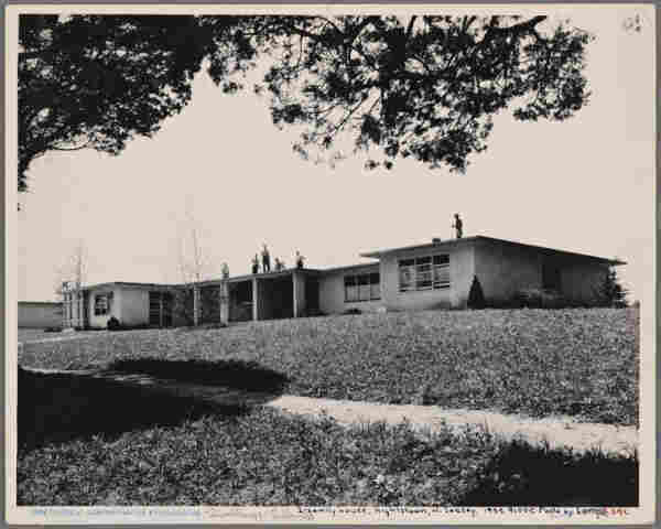  This is a black and white photograph depicting an older residential structure. The house features an L-shaped layout with multiple sections, possibly indicative of a two-family home. It's topped by flat roofs and has an unfinished appearance with visible exterior walls. There are trees in the foreground and what seems to be a clear sky in the background. The photograph includes text, which indicates that this house was completed in July 1936, suggesting that it may have been taken around that time or as part of historical documentation.