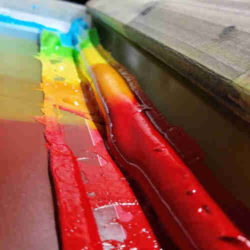 A close up of a wooden squeegee sitting in a screen, loaded with ink. The ink is in a rainbow gradient - blue farthest from the camera, then green, yellow, orange, and red. 