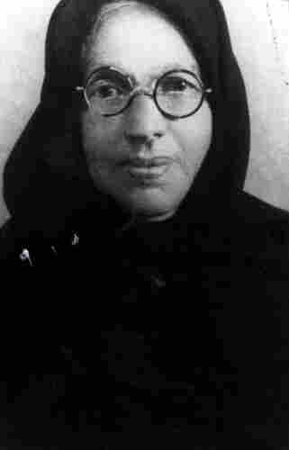 A photo of a women in round-framed glasses in dark clothes and a hood or scarf over her head.