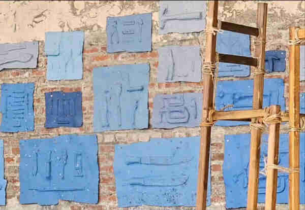 Blue cast paper relief sculptures of tools on a brick wall, partially obscured on the right by a wood and rope sculpture.