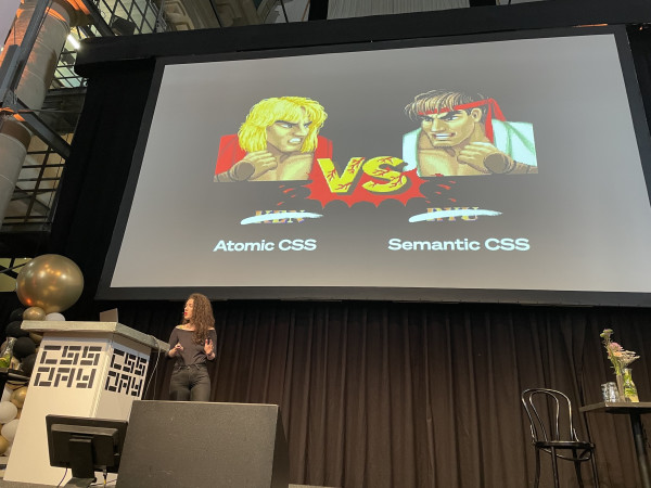 Sarah on stage with a slide of Ken vs Ryu, now Atomic CSS vs Semantic CSS