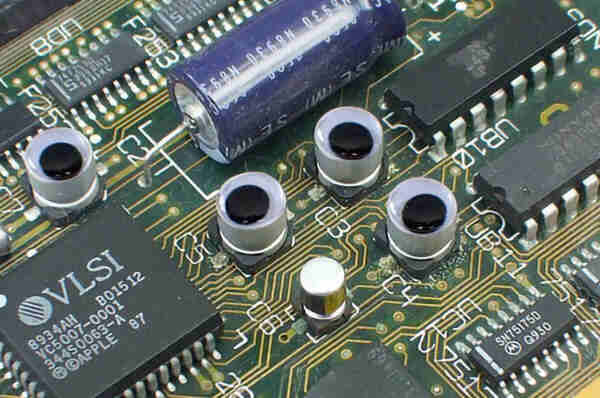 A photo of the closeup of a 68k Mac logic board with some chips, a large axial can capacitor, four capacitors with googly eyes on top, and one installed incorrectly without.