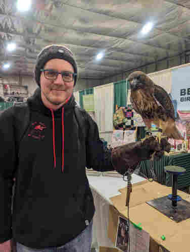 A photo of me in a toque and a black hoodie with a red Bak Mei Kung Fu logo on it. I've got a heavy falconry glove on my left hand, and on it is perched a red tailed hawk.

We're inside an arena set up with booths about birds and nature conservation, part of the annual Snow Goose Festival in Tofield, Alberta.