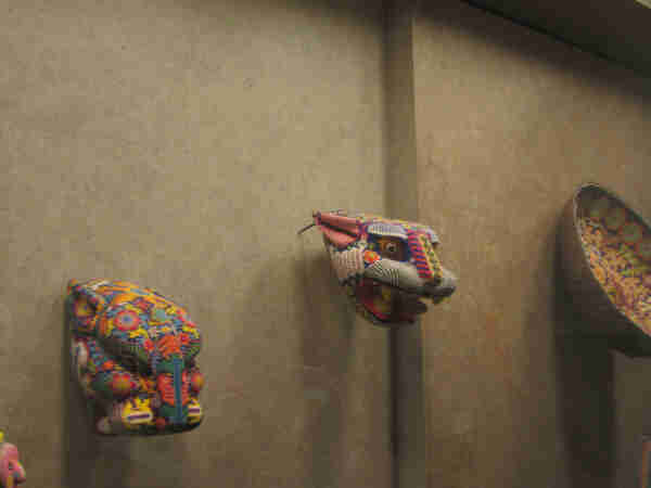2 wooden sculptures of animal heads and a basket displayed on a wall.  All have colorful beads arranged in different patterns placed on top of them. I believe a wax substance is placed on the wood and the beads are then pushed into the wax. A jaguar head is one of the animals.