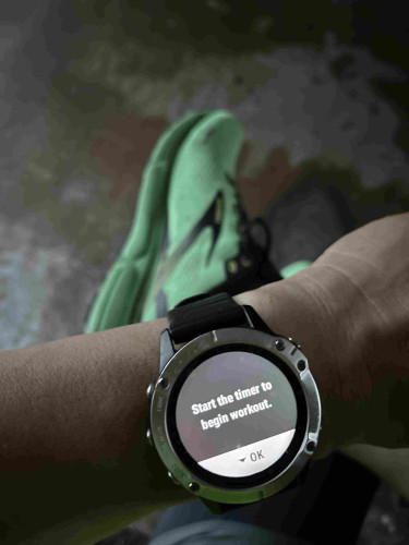 A photo featuring a person's wrist wearing a sports watch with the text "Start the timer to begin workout" and a blurry green running shoe in the background.