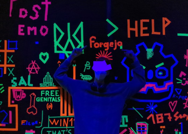The author of this post in front of a wall with tape of various shapes and drawings made by various people. The room is being lit by UV lighting, and the tape shines under that said UV lighting.

In the picture, I am pointing to a drawing (made, like everything else on this black wall, using that said tape), that looks like the Forgejo logo.

Other "drawings" can be seen all over the place. The logo of the Godot Game Engine, "Lost Emo", a small bug, tic-tac-toe, hearts and cat drawings, as well as, well, a phallic drawing that I only noticed in retrospect. I should have picked a better place to put the Forgejo logo, but I did not expect to share pictures here!

Photo taken by @linus@donotsta.re
