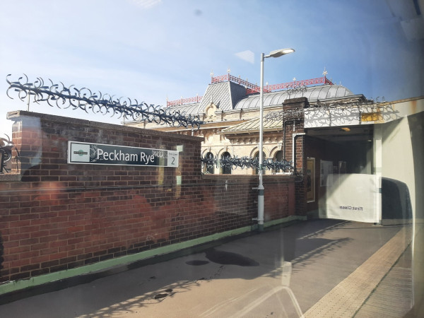 Photo shows a station name plate on a wall topped with rotating metal pointy objects. The platform recedes left to right. An ornate station roof is seen in the background.