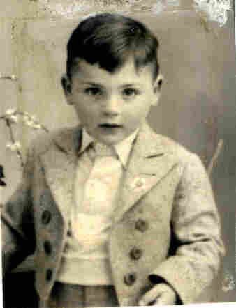 Studio photograph of a young boy photographed against a solid background. He has dark short hair. His fringe falls over his forehead. He is wearing a jacket with three buttons - unbuttoned, a waistcoat and a shirt buttoned up to the neck.