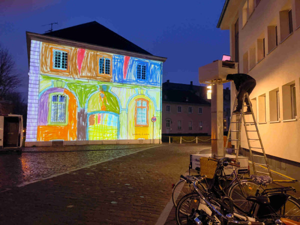 A night view of a house onto which a colorful children’s drawing of the same house is being projected. On the right a man stands on a step ladder in front of the large projector, in front of a row of parked bicycles. The cobblestone street leads off to the right, the dark blue late evening sky is clear and empty. 