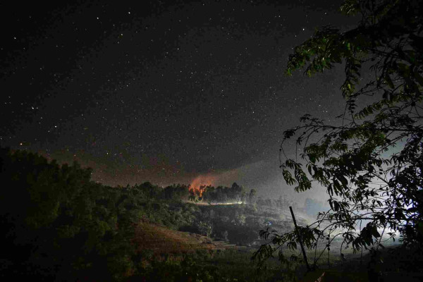 Smoke rising behind the closest hill lit up orange by fires underneath. Starry sky above.