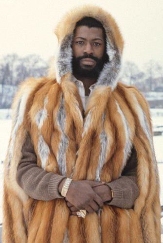 A portrait of Teddy Pendergrass taken in 1979 by the photographer Bernard Gotfryd. Teddy looks dead on at the camera with his hands clasped loosely in front of his waist. He is outside on a bright, snowy day, wearing a hooded fox fur coat. His gaze is distant but penetrating. He wears a gold bracelet that says "TEDDY," as well as hefty gold and diamond rings on his little fingers.