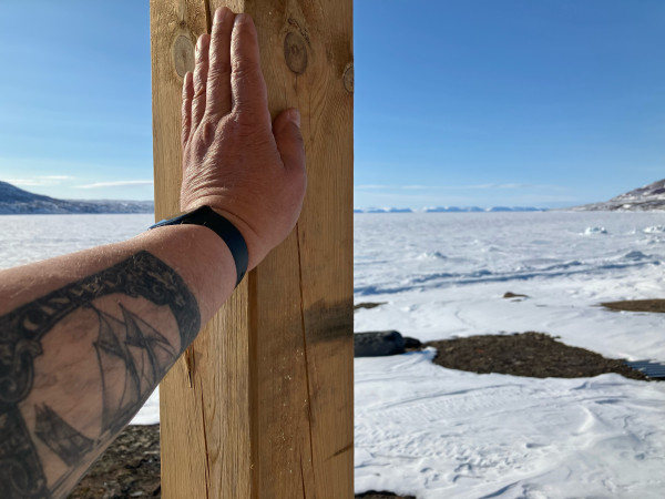 An arm with a tattoo of a 1929 Canada 50¢ Bluenose stamp leans up against a post. The frozen Arctic Ocean stretches off to a far away line of cliffs, surmounted by clear blue skies. 