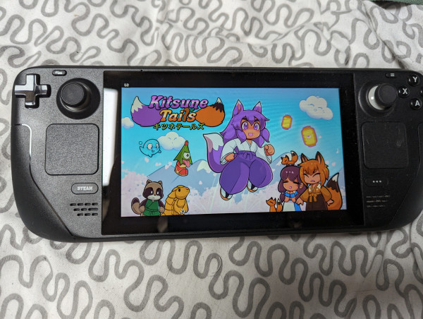 A  Steam Deck shows the Kitsune Tails title screen. A purple-haired fox girl in a white shirt and purple trousers leaps into the sky. Other on screen characters, further in the background, include a red fox-girl a human with a red box on her head, a tanuki wearing a green robe, and a selection of cute monsters, including a one-footed hopping triangular yokai, maybe a water sprite and a pangolin-themed thing.