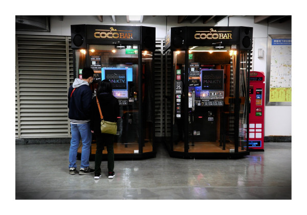 The image is a color photograph taken in Taipei, showing two individuals standing in front of two adjacent kiosks labeled "The COCO BAR." These kiosks are designed for private karaoke sessions, as indicated by the "MiNi K.T.V." signage on the screens inside each kiosk. The individuals, seen from behind, appear to be examining the kiosks, possibly preparing to use them. The surrounding area looks like part of an indoor public space or a transit station, with a map and a change machine visible on the right side of the image. The overall atmosphere suggests a modern urban setting.