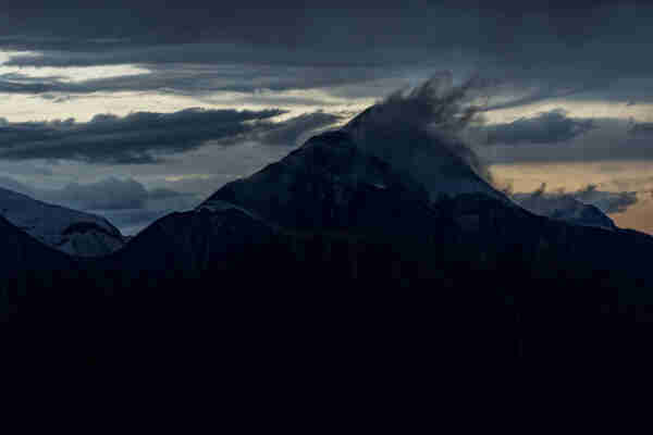 A snow-capped mountain, dark at sunset, is flagged by mist and wind at its peak; in the background, torn and stormy clouds furrow the sky in shades of steel grey, split with palest gold, concentrating to pale orange on the right. 