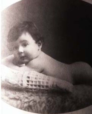 Vintage monochrome photograph of a baby boy reclining on a cushion, looking directly at the camera.