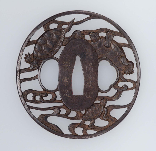 official museum photo of the object, front view on light grey background: iron tsuba (Japanese sword guard) depicting three turtles swimming in waves around central hole