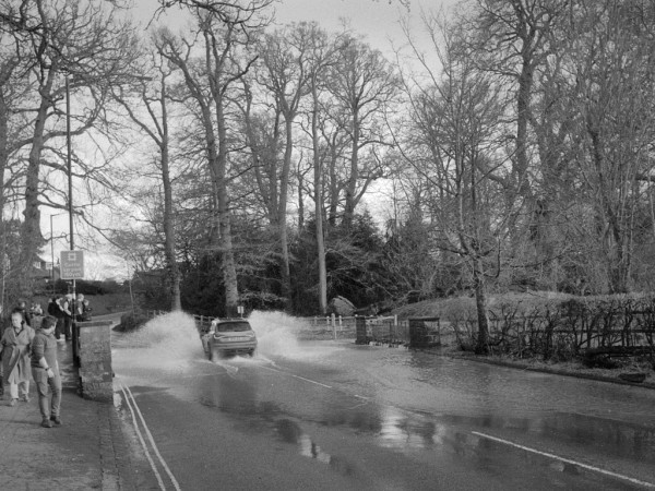 A car drives through the local ford when it has flooded, raising a splash higher than the car's roof. A group of locals stand at the railings on the left, while a young man walking towards the camera turns to look back. Bare winter trees behind. Black and white photo.
