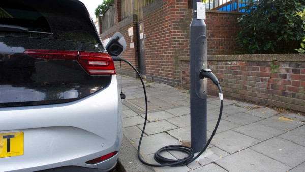 A white electric car is plugged into a lamppost charging station located on a sidewalk next to a brick wall.