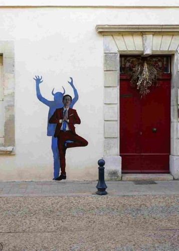 Streetartwall. A self-designed, life-size paste-up of a young man in a yoga pose is stuck to a beige exterior wall between a window and a red wooden door. The dark-haired man in a wine-red suit, white shirt and blue tie is standing on one leg and has his eyes closed. He has folded his hands and appears to be meditating. His shadow behind him reveals what it looks like inside him, for there is an agitated dark devil standing on one leg and waving his arms.
Info: The artist Charles Leval, alias Levalet, stands out from the usual forms of street art, as his scenes, lovingly drawn with ink on paper, show figures in their (often tragic-comic) everyday lives. They seem like repressed, unconscious feelings and experiences. They often fill long walls and are even told as serial stories.
For the "Les Recontres d' Aubergine" festival, he distributed some pasteups around the city.