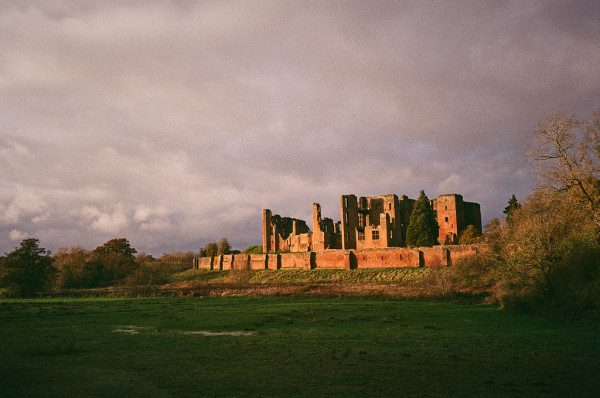 Photo of Kenilworth Castle in late afternoon autumn light, with a cloudy, slightly yellowed sky above. The foreground grass is in shadow, but still shows a sliver of water from one of the many floods we've had in the past 6 months.