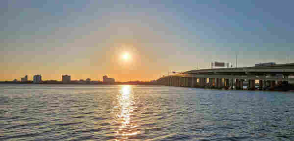 A golden glow at the horizon highlights a city skyline as the sun lowers in a clear blue sky over a wavy blue river, right next to a major interstate highway bridge crossing the wide river.