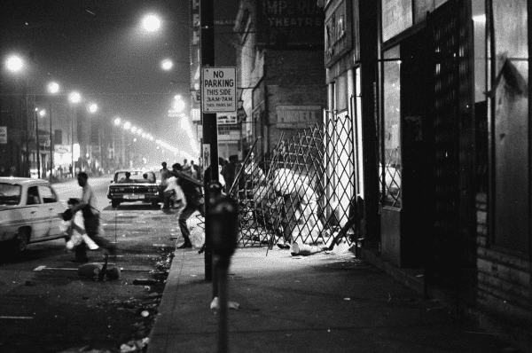 Along a dark city street at night, people appear to be fleeing a scene of disarray.