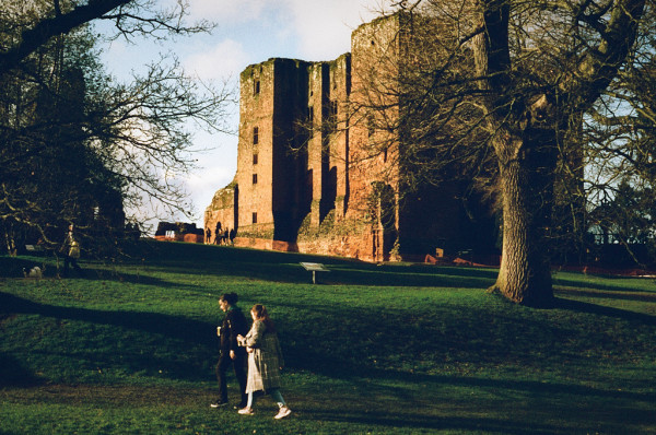 A couple walk across the grass in front of the massive keep of Kenilworth Castle, which is lit by the afternoon sun.