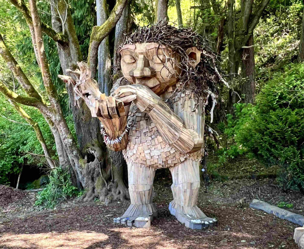 A giant but gentle wooden troll is playing a large wooden flute. Her eyes are closed in reverie. She has long, twiggy hair and a beach bracelet on her right wrist. Offerings of flowers and shells have been left on her toes. Background is a very green foresty forest!