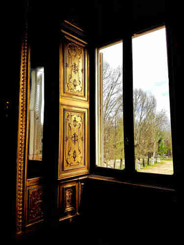 An ancient window, from the inside. It's made of wood, finely crafted, ready to be closed to cover the daylight. The light coming from outside gives it a golden, noble appearance. It's from the Royal Palace of Turin, an important window. Outside, some bare trees, a street, some statues.
There's also a mirror, next to it, ready to reflect the light and show, from the inside, what's happening outside