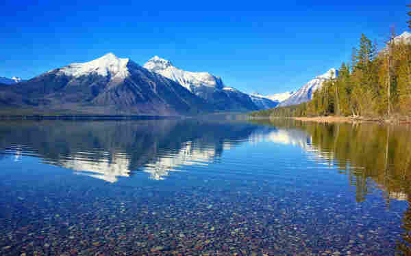 Lake McDonald, Glacier National Park, USA. Large calm lake with clear water. We can see smooth stones coating the bottom of the lake in the foreground. Farther away, the lake reflects snow-capped mountains in the distance. Sky above is clear and blue. Some evergreen trees are seen along the side of the lake.