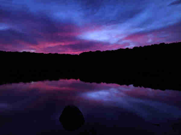 It is well past sunset with a deep blue twilight settled over the scene. Clouds near the western horizon are still tinted vibrant dark pink and deep purple shades, mirrored on the very slightly rippled lake surface below. A partially submerged rock a few feet offshore and the entire wooded far shore of the lake are silhouetted black in the dim light.