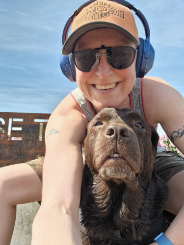 Selfie of Arwen and myself. Arwen sits in front of me, her mouth slightly open (some tootsies are visible). My head is above hers, I'm smiling at the camera. Wearing a orange Harley-Davidson cap, blue headphones and black sunglasses. 