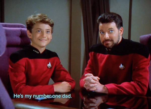 TNG scene. Hallmark channel screenshot vibes here. We're on the D and in the conference room and Riker is shown sitting at the table, hands clasped, looking up with a half smile. To his left, also sitting, w/ arms on the table, is a smiling and happy looking young man. He's maybe 13 or 14 years old, and wearing the same red uniform and comm badge as Riker. Caption is centered over the little guy, and reads, "He's my number-one dad."
